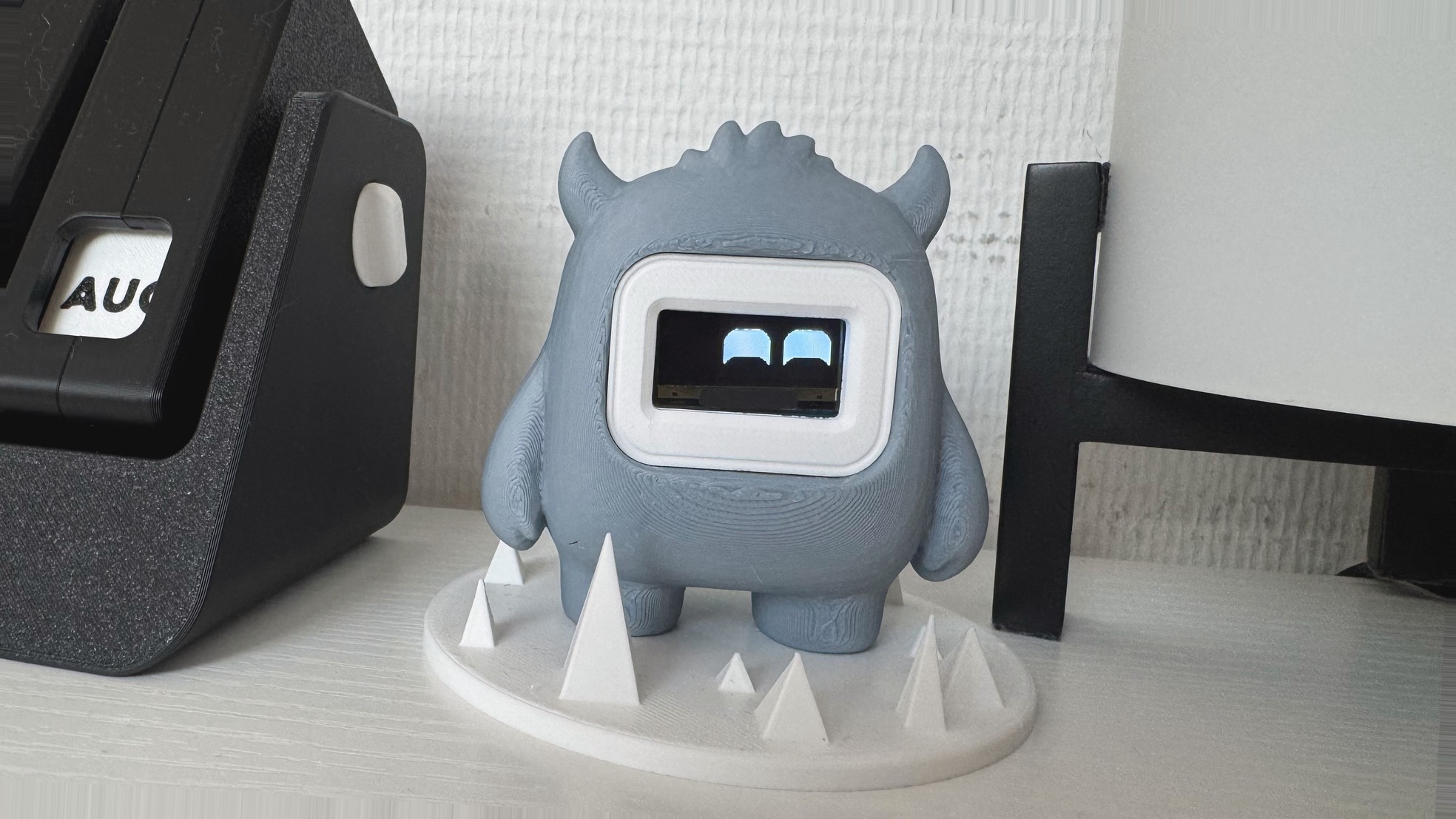 Blip on the desk: a grey 3D-printed creature with two horns, a white bezel and two arched blue eyes, standing on a white spiked base. An August calendar sits to the left.