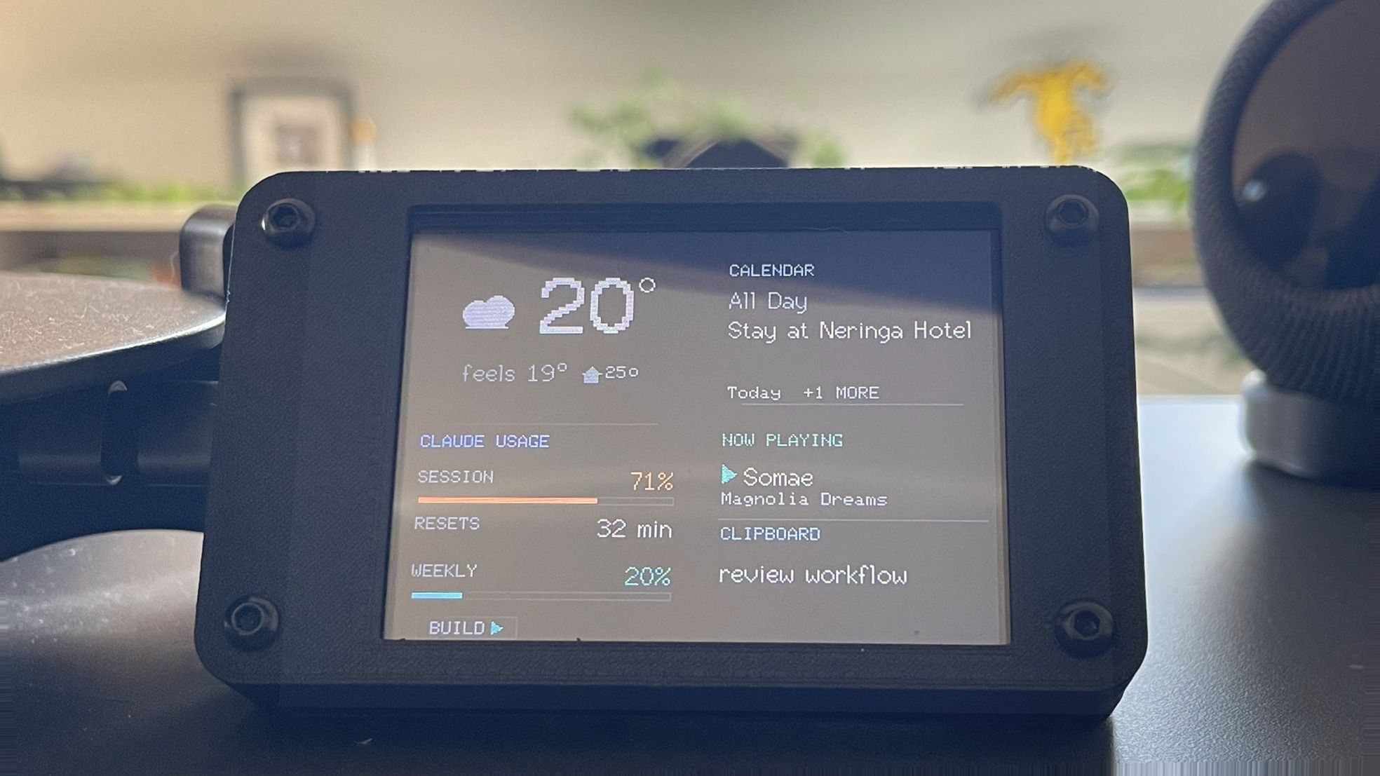 The landscape glance screen on the desk: 20 degrees partly cloudy, the calendar reading Stay at Neringa Hotel, Claude session at 71 percent resetting in 32 min, weekly 20 percent, now playing Somae, and the clipboard reading review workflow.