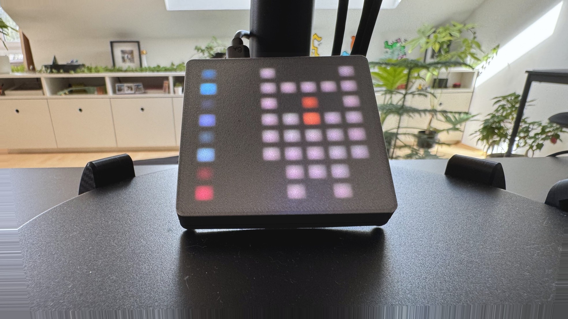 Eight on the shed desk: a charcoal 8x8 LED panel showing a pale owl face with orange-red pixels and a left status rail, plants and white cabinets behind.